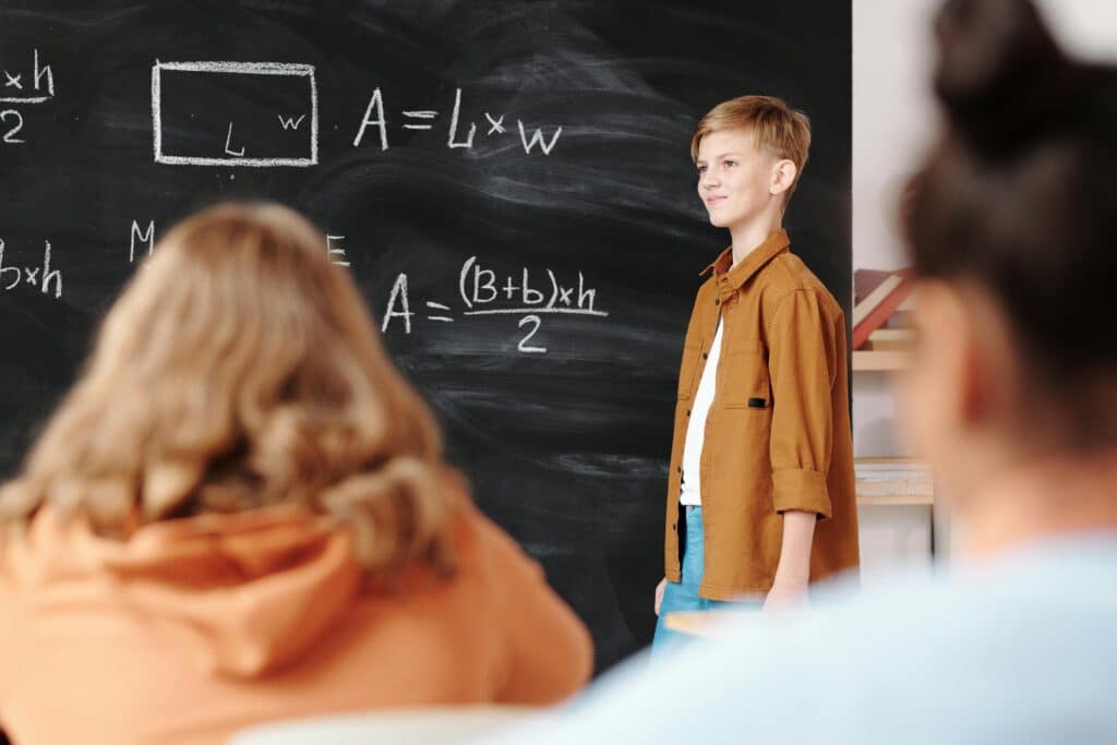A young student confidently presenting math formulas on a blackboard in a classroom setting.