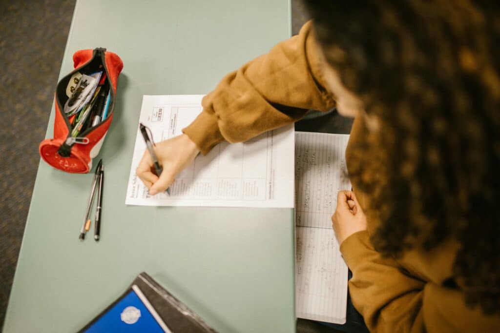 Student writing on exam paper at desk during class with notes and pencil case.
