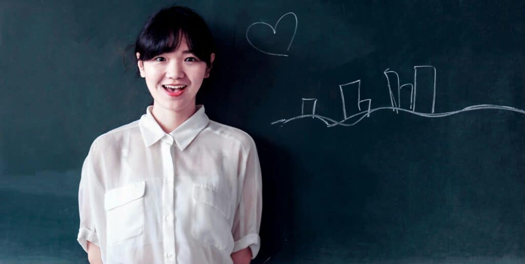 Smiling young woman stands confidently by a blackboard with chalk drawings.