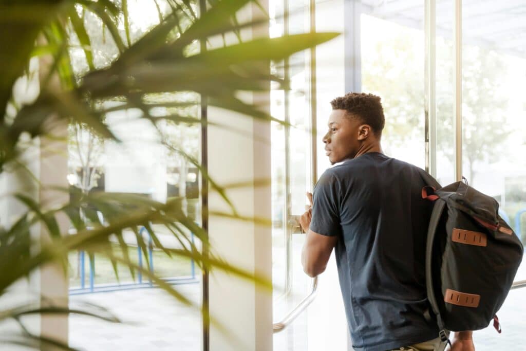 A college student with a backpack exits a campus building through glass doors.