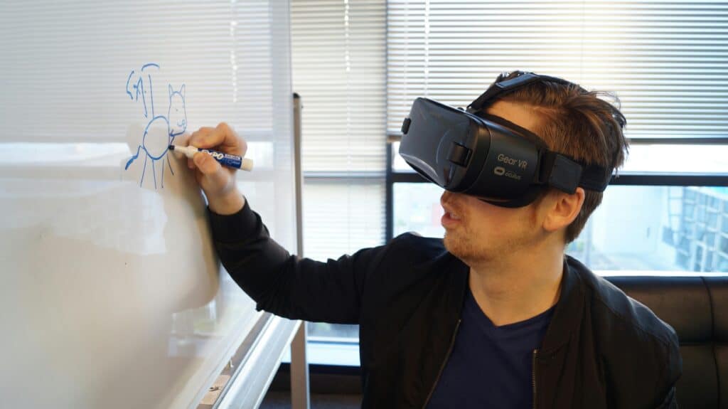 A man wearing a VR headset drawing a cat on a whiteboard in an office environment.