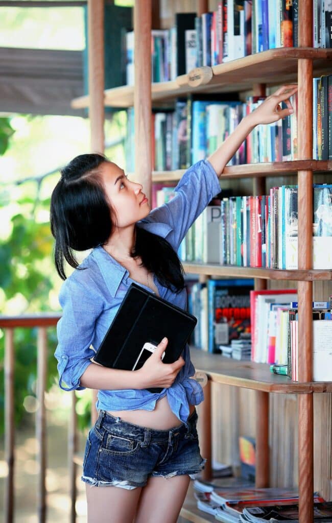 A young woman reaches for a book on a library shelf, holding a tablet.