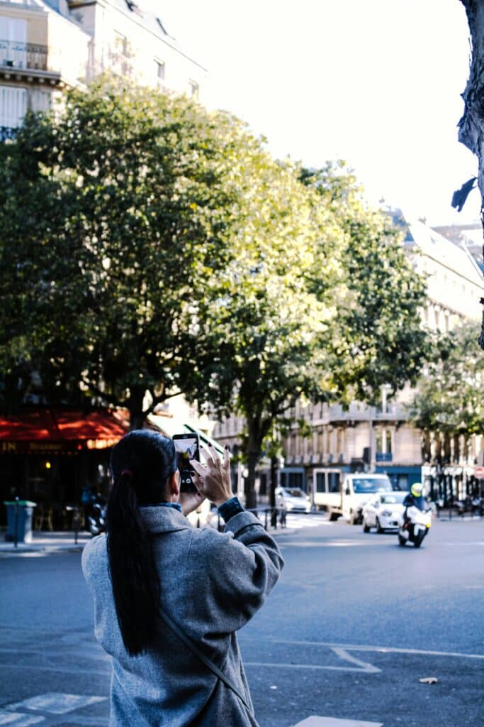 A candid street scene in Paris showing a woman taking a photo with her smartphone. Captured on a sunny day, the image highlights the charm of Parisian architecture, leafy trees, and everyd...