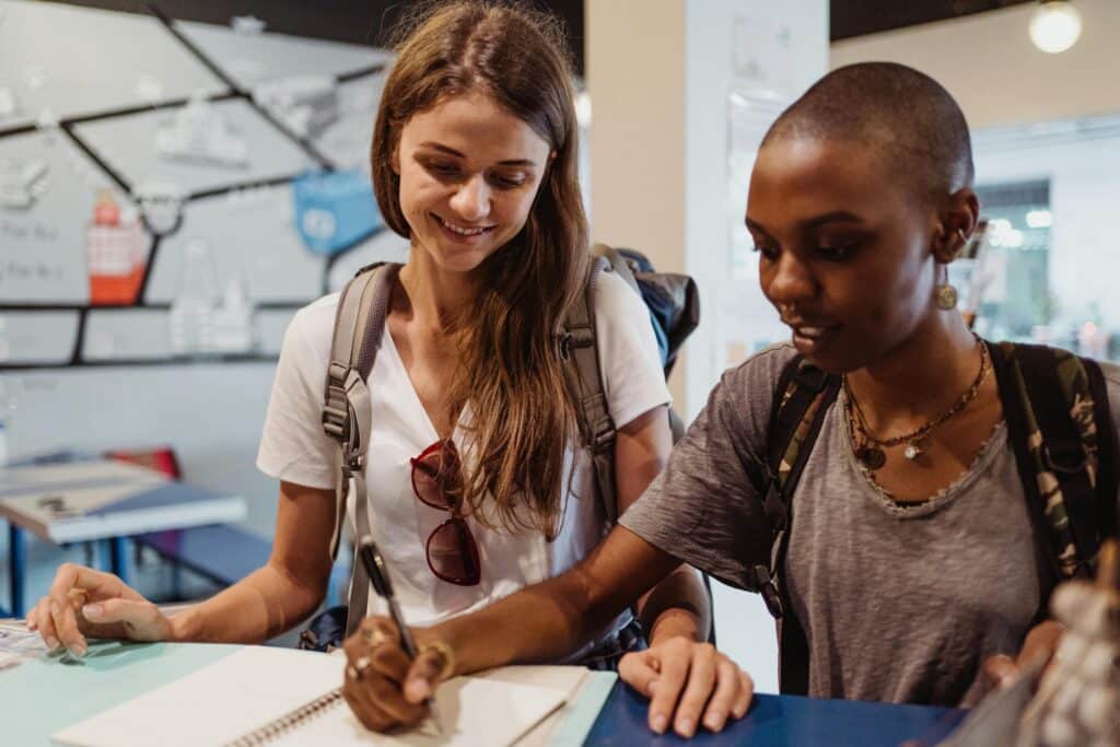 Two women with backpacks signing a guestbook in a hostel, smiling and engaged.
