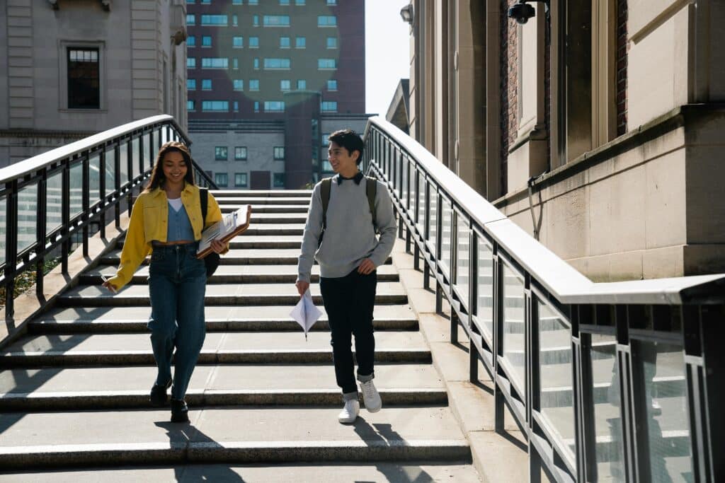 Two university students walking down a staircase, carrying documents and smiling during the day.