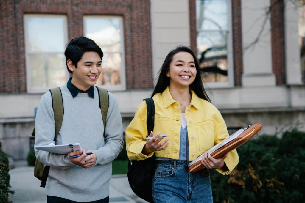 Two smiling students walking outdoors on a college campus, carrying supplies.