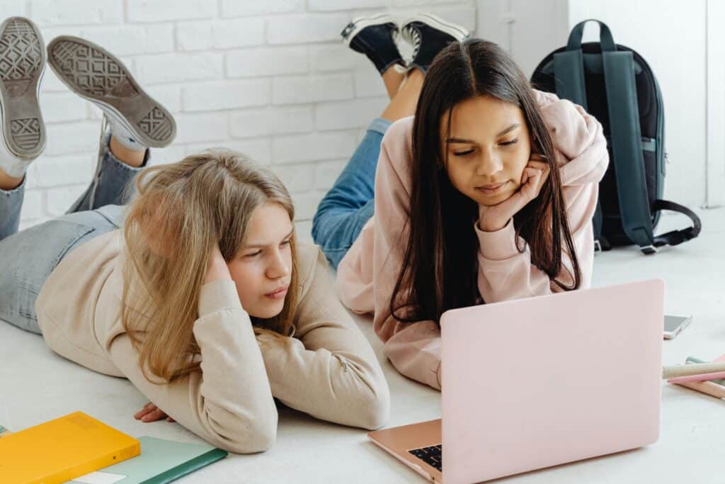 Two teenage girls studying together using a laptop indoors, emphasizing friendship and education.