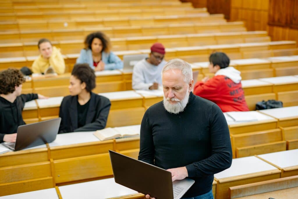 Mature teacher interacting with diverse students using laptops in a spacious lecture hall setting.
