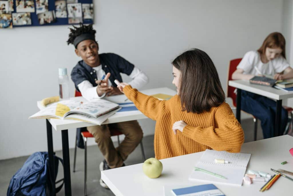 Students interacting in a classroom with textbooks and school supplies on desks.