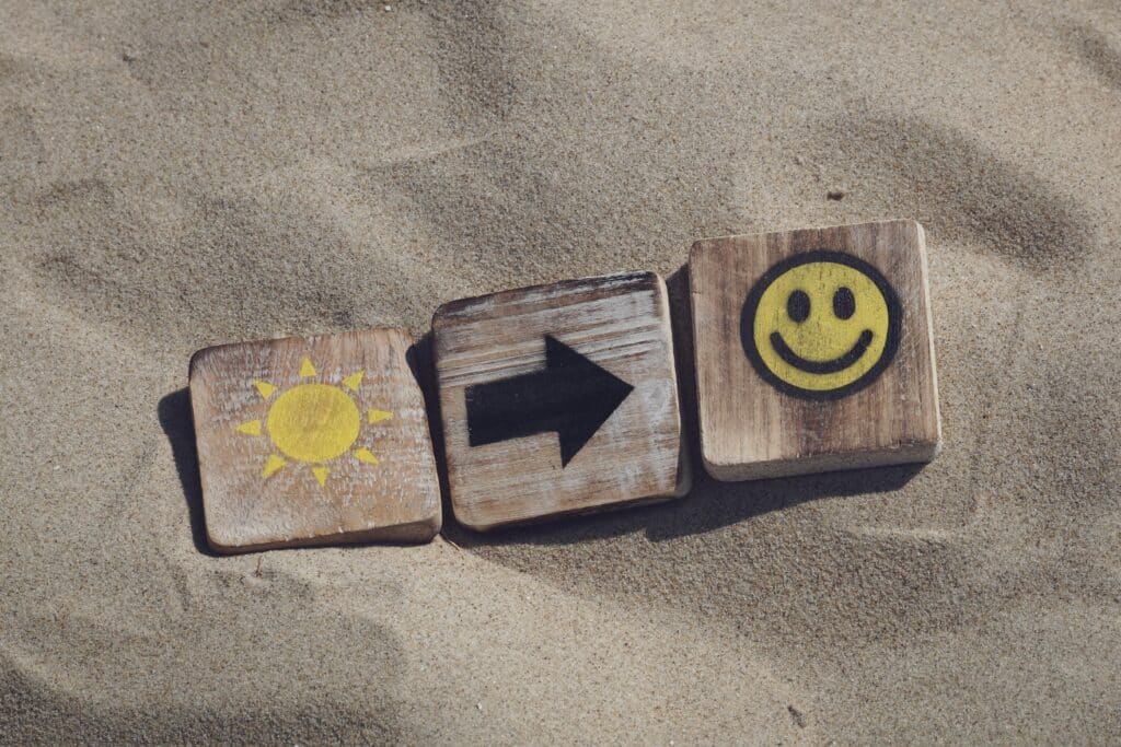Wooden blocks with sun, arrow, and smiley emoticons on sand, evoking summer vibes.