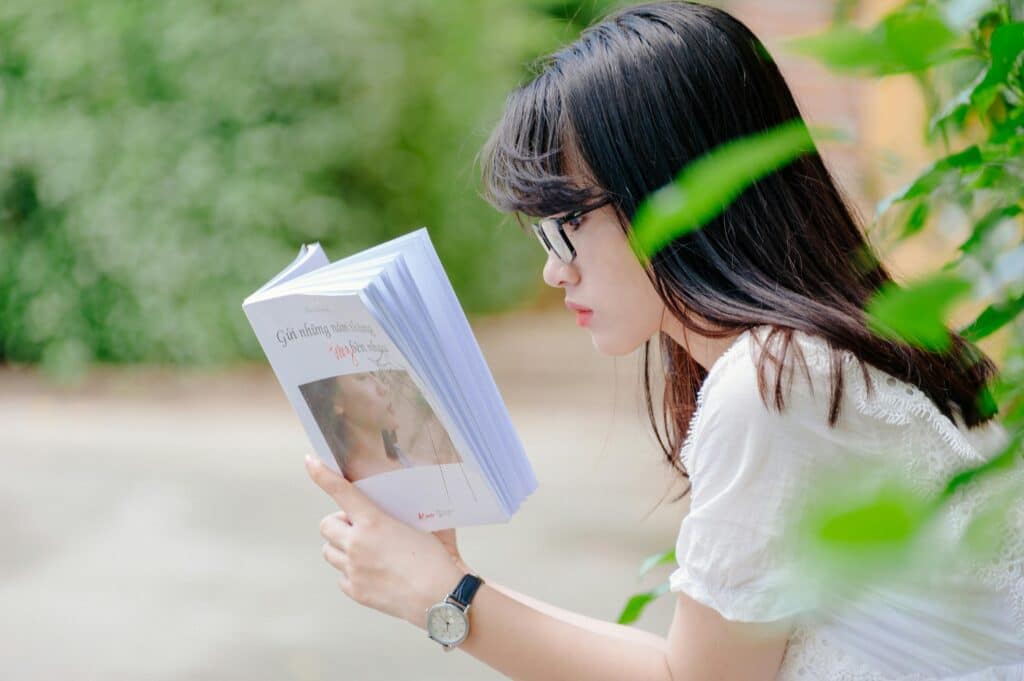 A young woman sits outdoors, absorbed in reading a book during a sunny summer day.