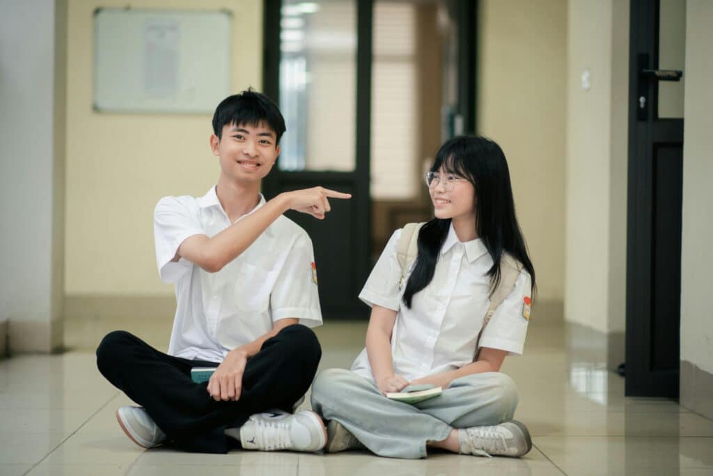 Two high school students sit laughing on the floor in a hallway, enjoying a playful moment together.