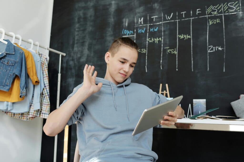 A teenager uses a tablet for online learning in a cozy home setting, waving at the screen.