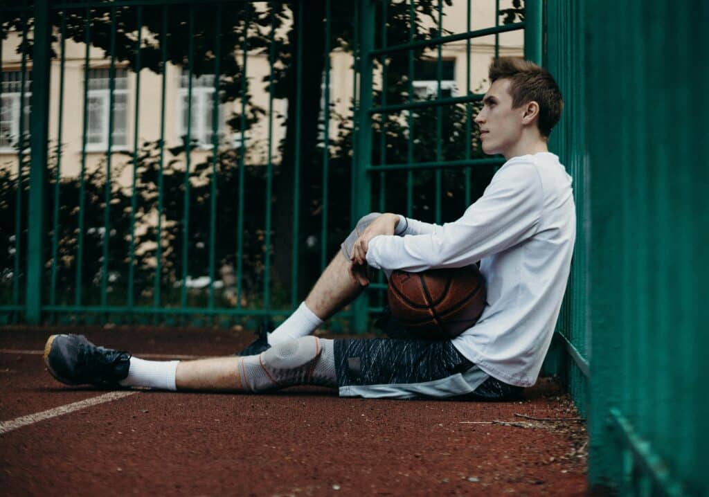 A young man sits with a basketball, taking a break on a fenced court.