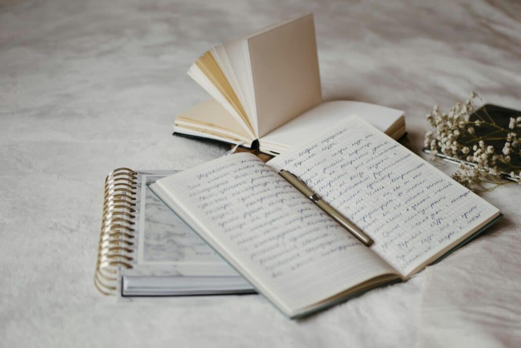 Close-up of open notebooks and a pen on a soft bed surface with delicate dried flowers.