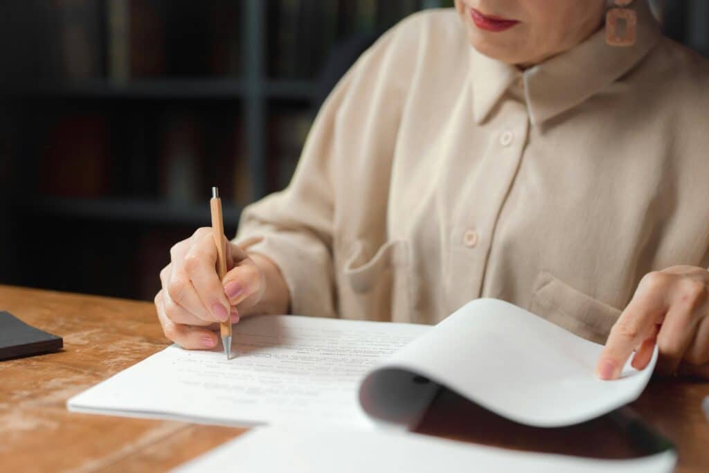 A woman reviewing and writing on documents at a wooden desk in an indoor setting.