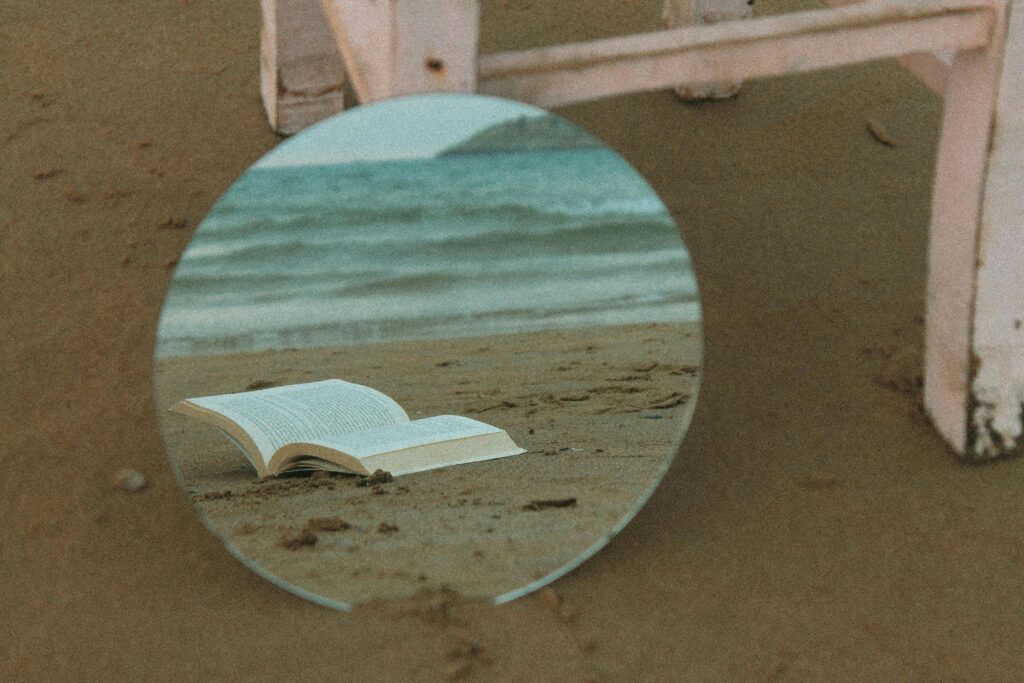 A round mirror reflects an open book on a sandy beach with waves in the background.