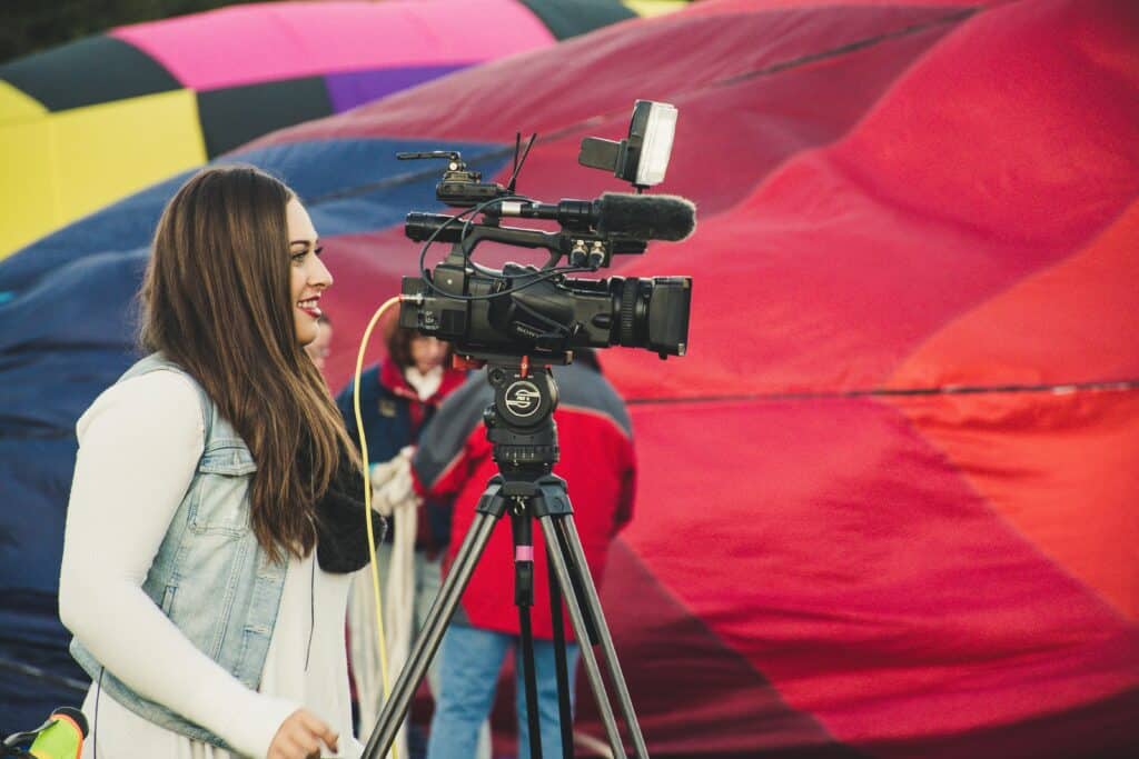 A female videographer operates a camera before a colorful hot air balloon backdrop.