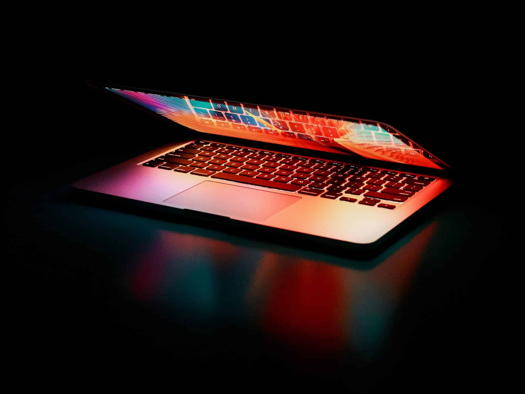 Laptop partially opened, emitting vibrant multicolored lights in dark setting.