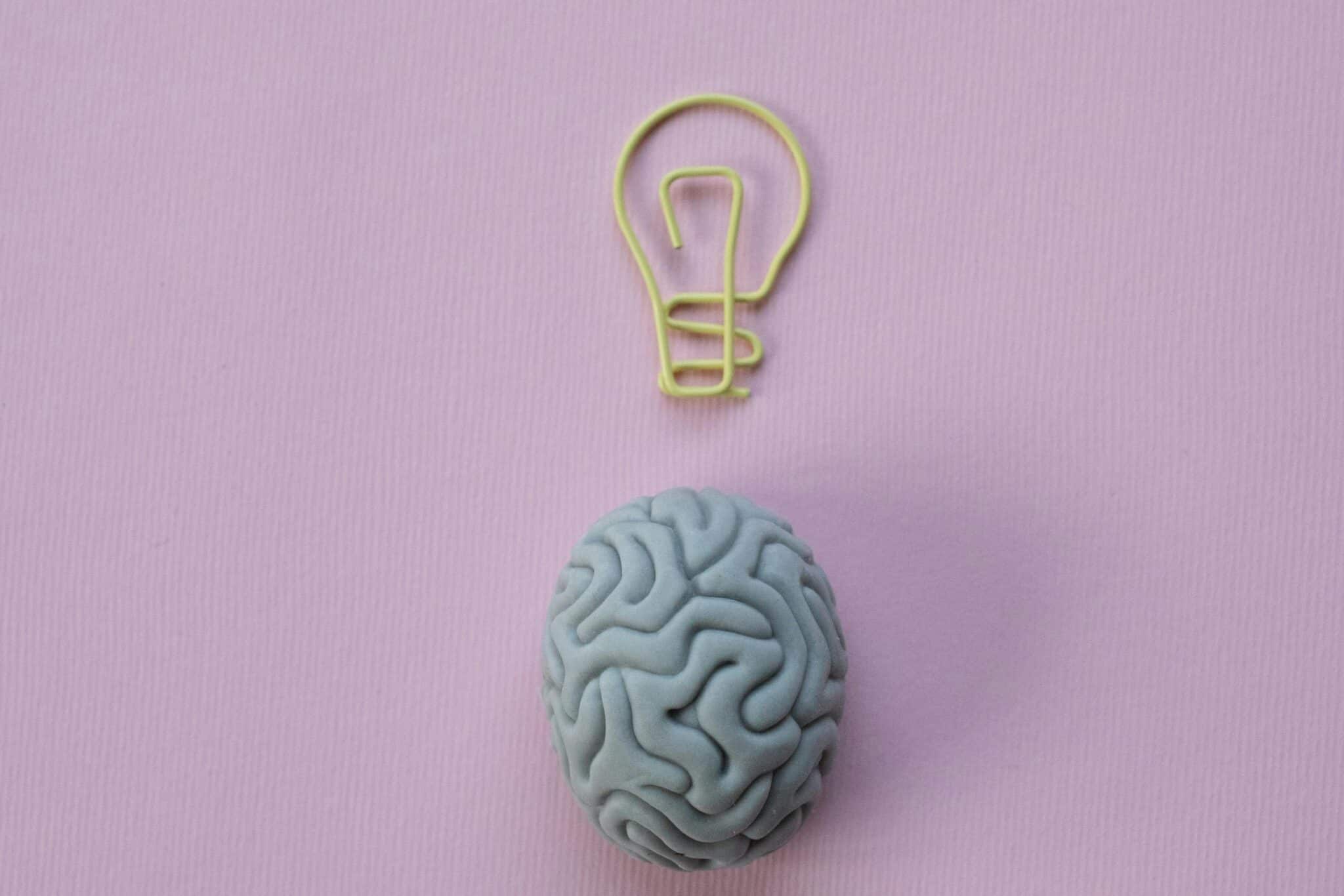 Brain-shaped eraser with a light bulb paperclip on a pastel pink background. Ideal for education and creativity themes.