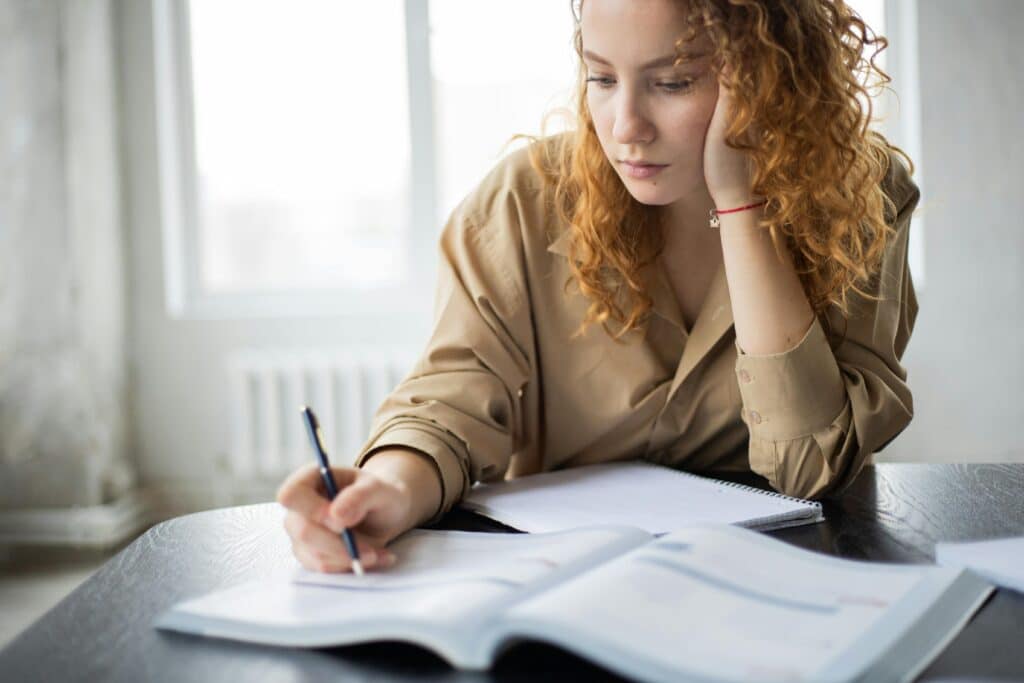 Woman with curly hair deeply focused on reading and writing at a desk.