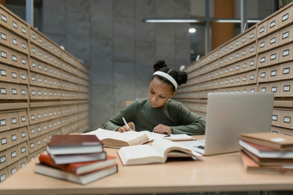 Dedicated African American girl studying in a library with books and a laptop.