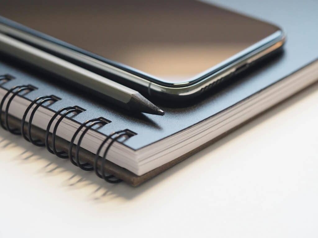 Close-up of a spiral notebook, smartphone, and pencil on a desk, highlighting modern workspace essentials.