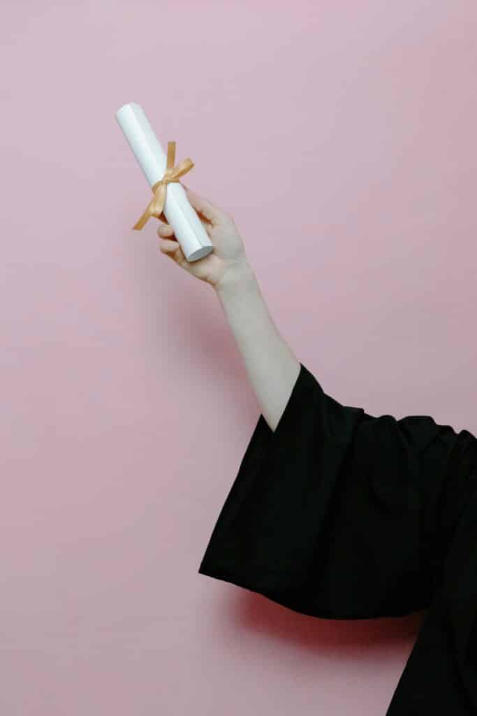 Hand of a graduate wearing a gown holding a diploma with a pink background.