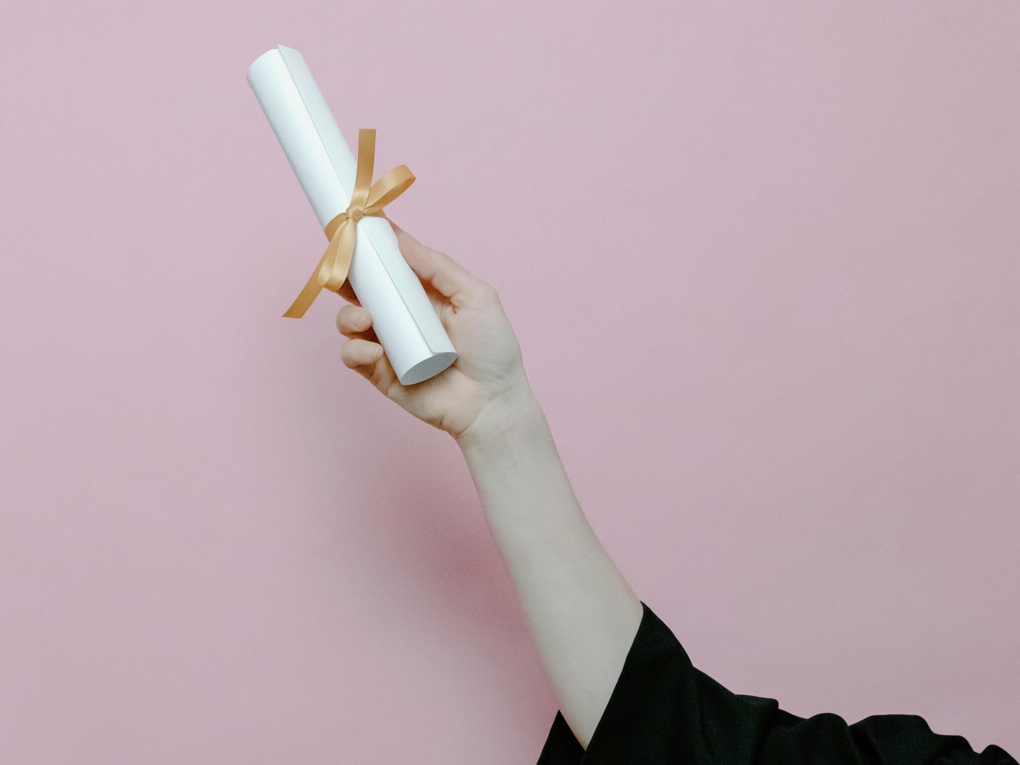 Hand of a graduate wearing a gown holding a diploma with a pink background.