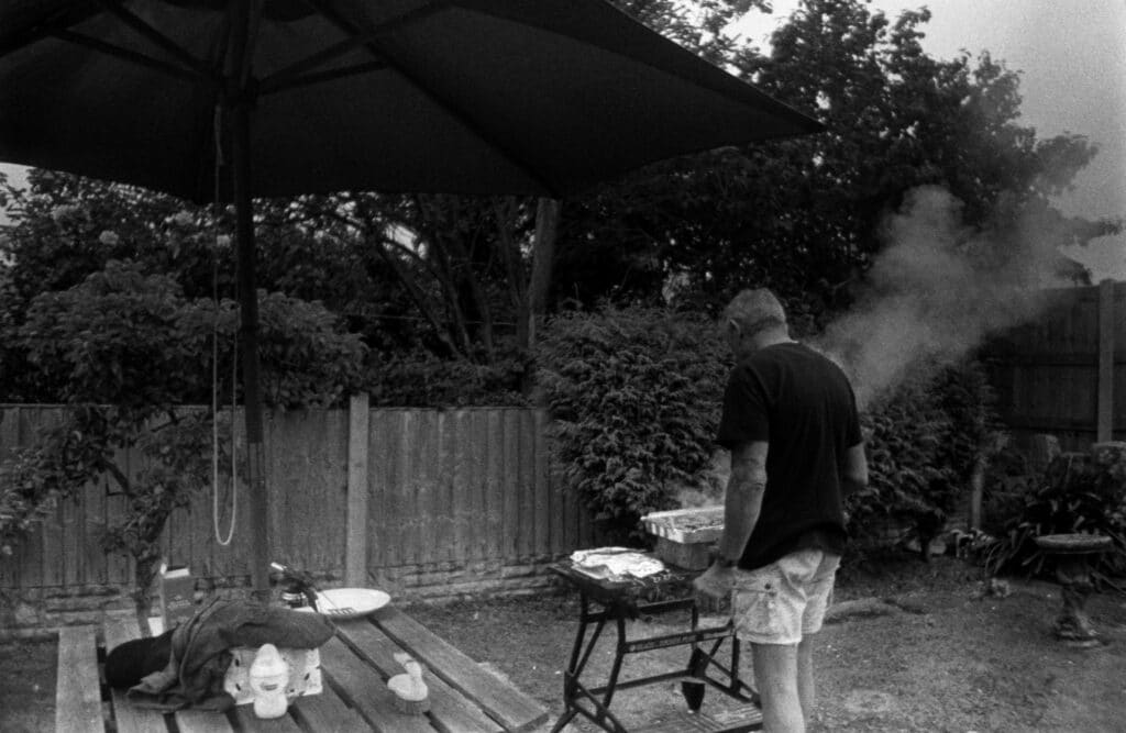 A man grills in a backyard barbecue scene under a patio umbrella.