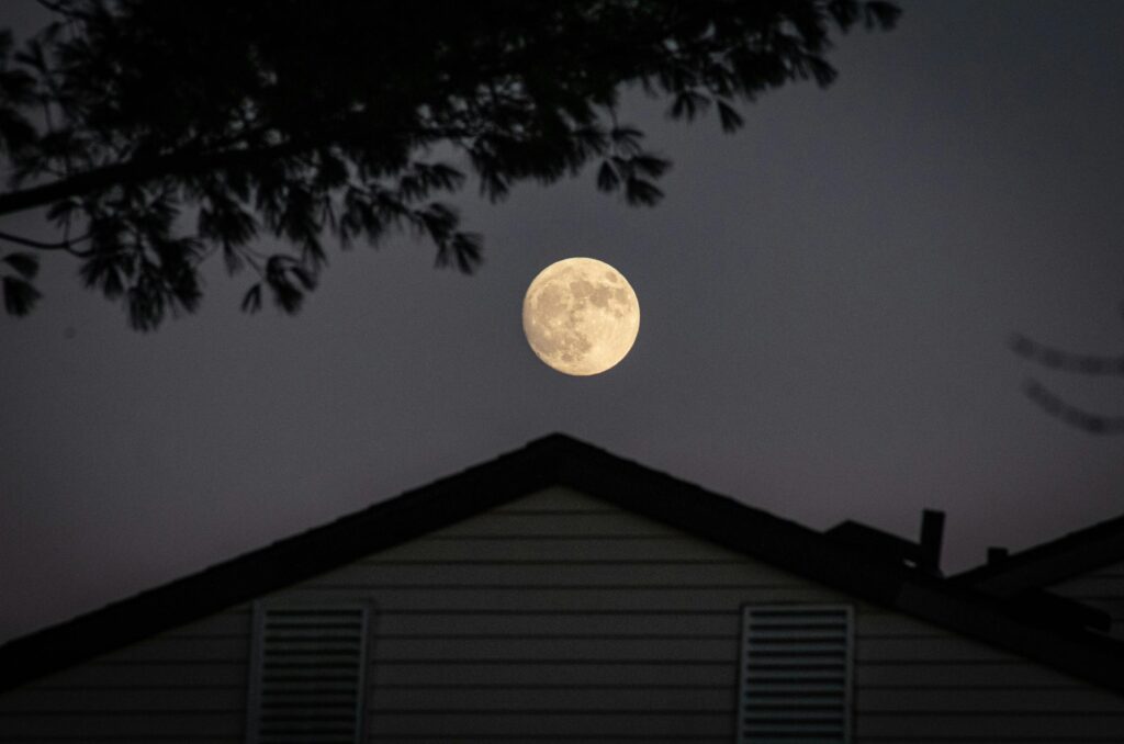 A full moon illuminating the night sky above a silhouetted house with trees.