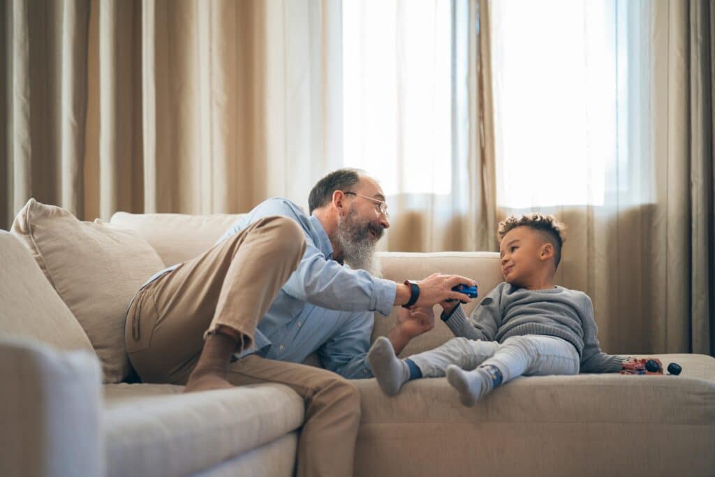 A heartwarming moment between grandfather and grandson laughing and playing on a cozy sofa indoors.