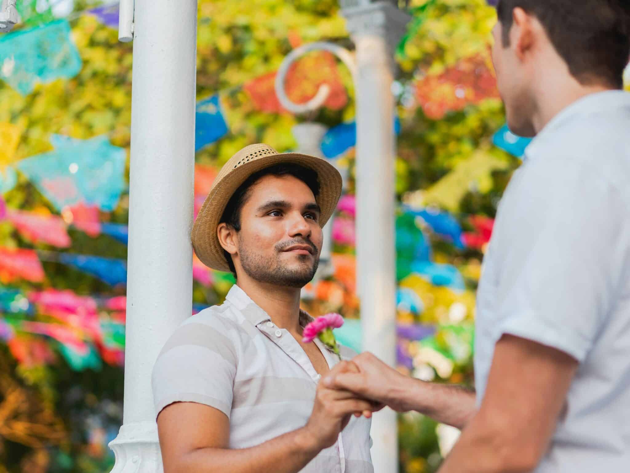 A gay couple holding hands outdoors, showcasing love and romance amid colorful decorations.