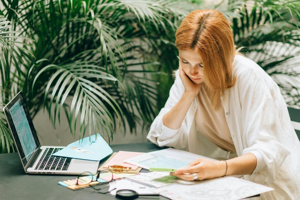 Woman in a thoughtful pose reviewing documents on a table with a laptop, surrounded by greenery.