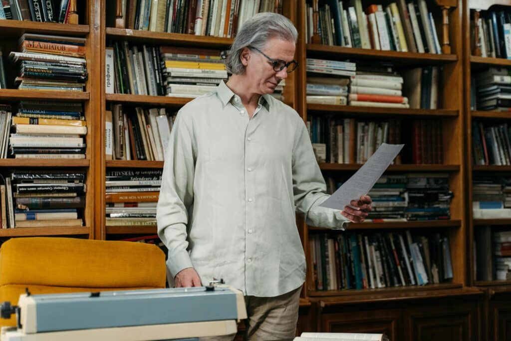 Elderly man reading paper in a cozy library with bookshelves and typewriter.