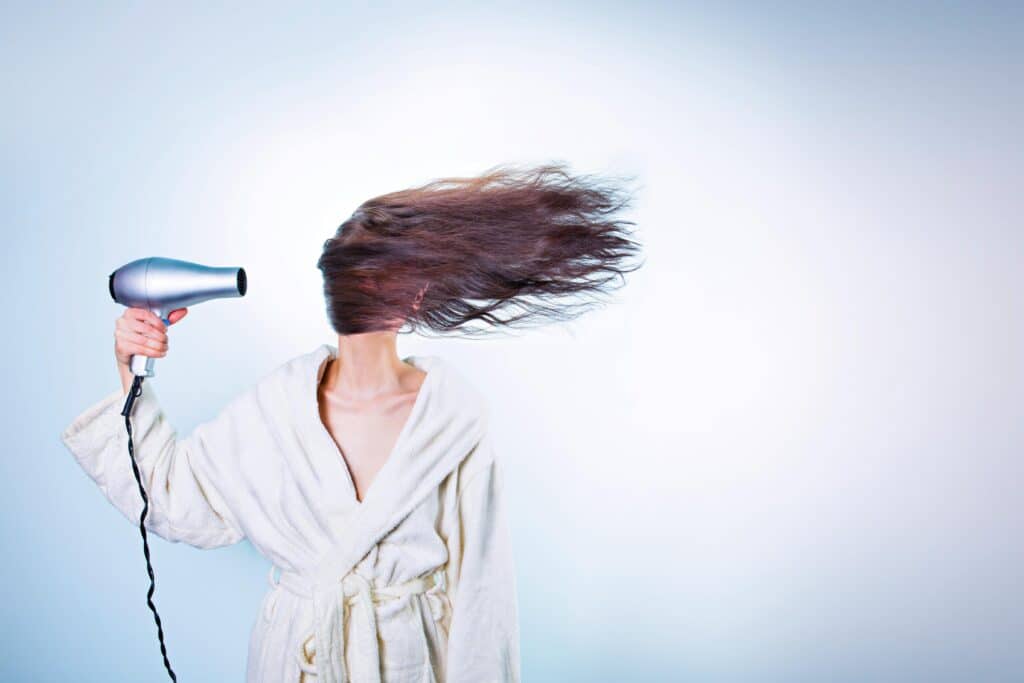 Woman in a bathrobe using a hairdryer, hair flying wildly. Indoor setting.