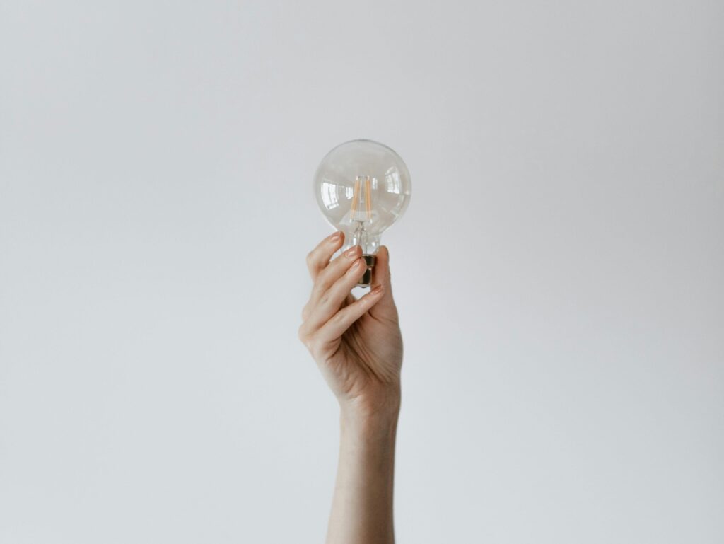 A hand raises a light bulb against a soft gray background, symbolizing creativity and ideas.