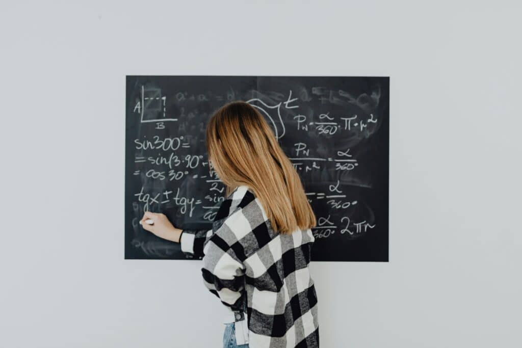 Teen girl in plaid shirt solving math equations on a blackboard, focusing on trigonometry.