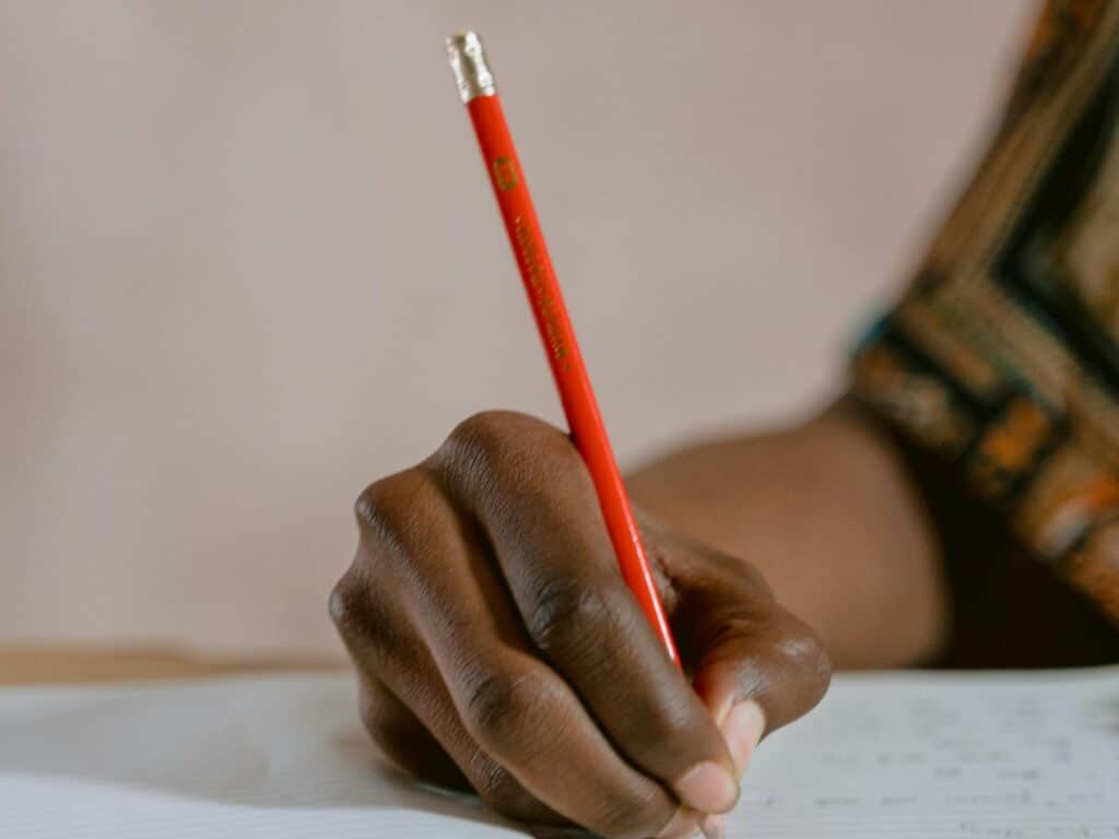 A person writing in a notebook with a red pencil, focusing on the hand's detail.