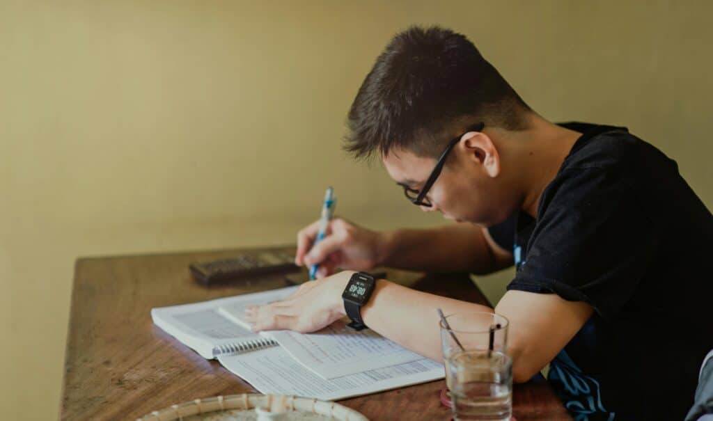 A young man concentrates on studying at his desk, taking notes indoors.