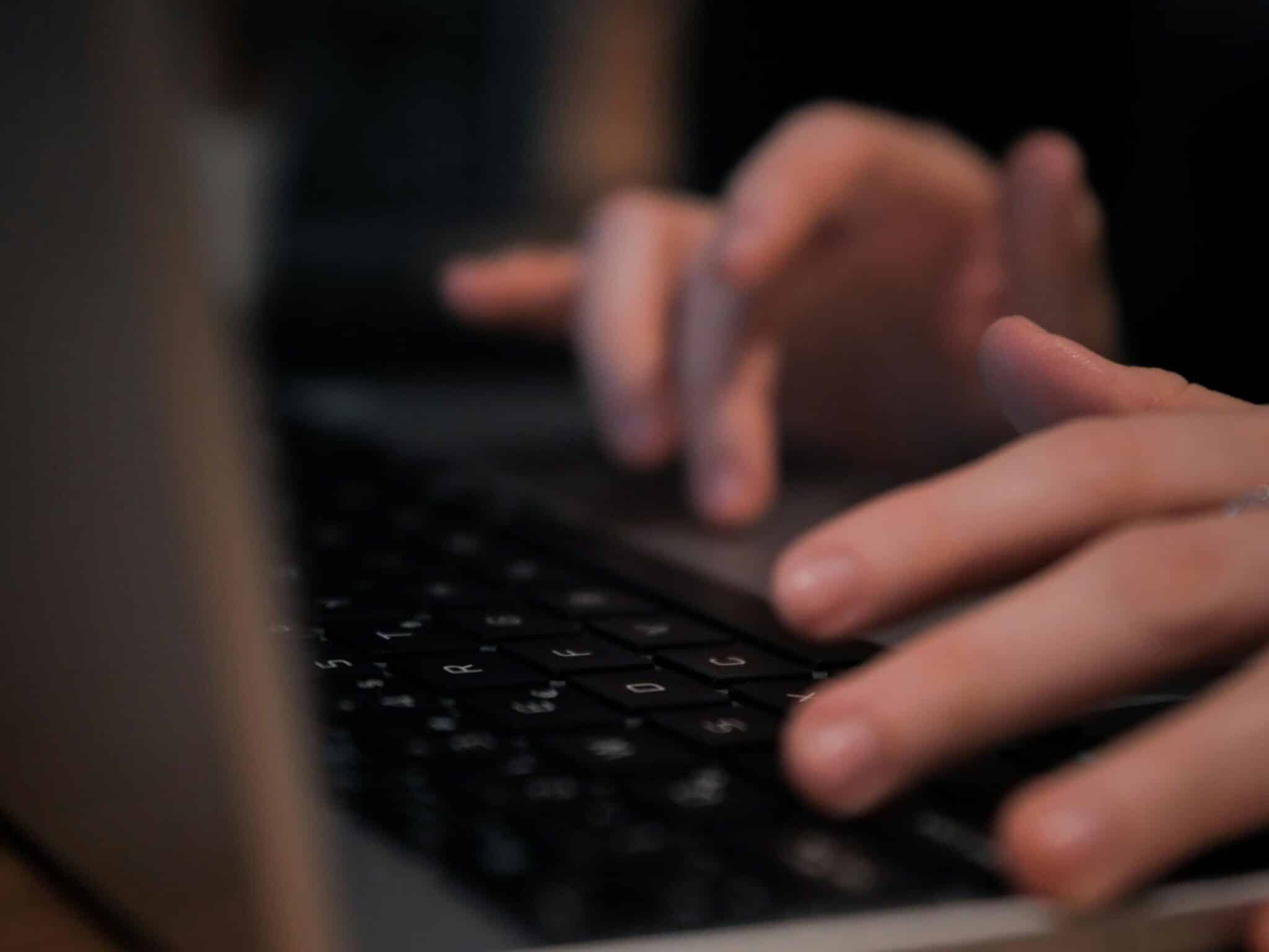 Blurred close-up shot of hands quickly typing on a laptop keyboard in a dimly lit setting.