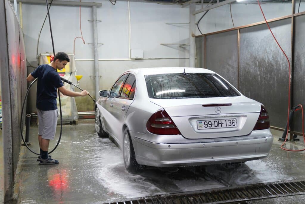 A young man using a hose to wash a silver car inside an indoor car wash facility.