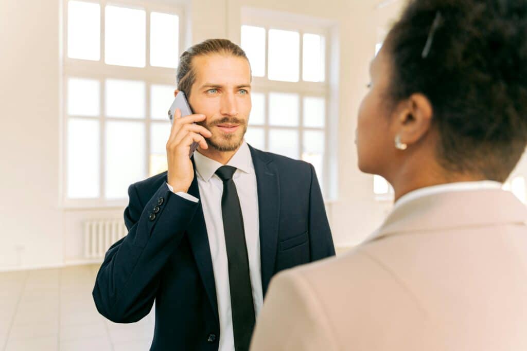 Man in business suit talking on phone with colleague in bright office setting.