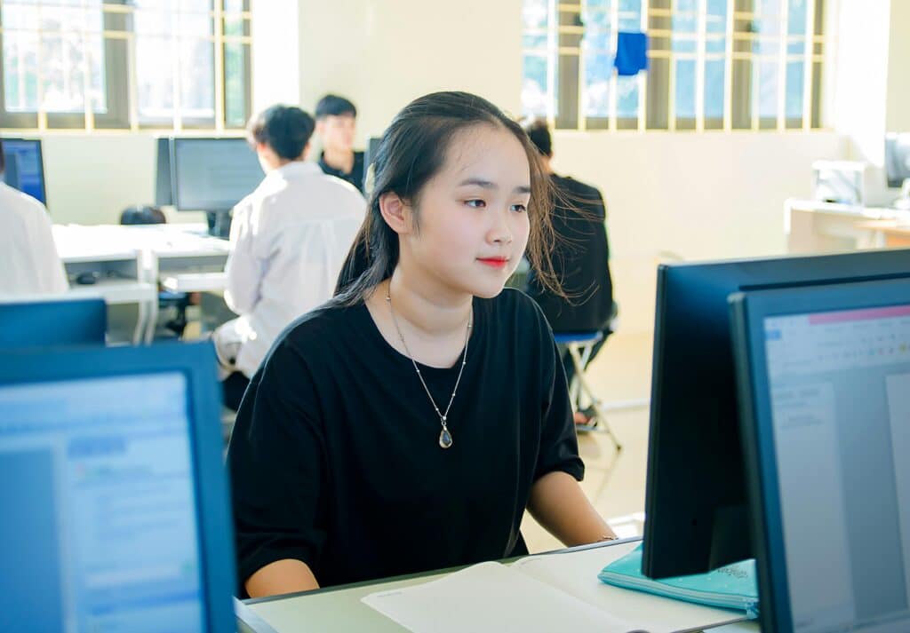 A young Asian woman concentrating on a computer screen in a university classroom.