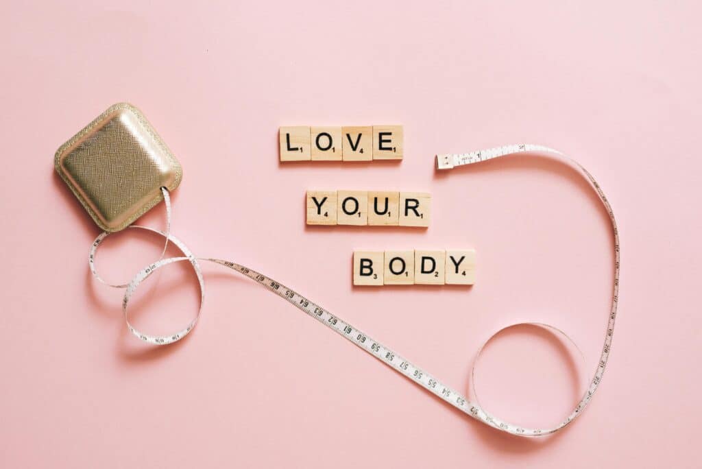 Scrabble letters spelling 'Love Your Body' with a tape measure on a pink backdrop.