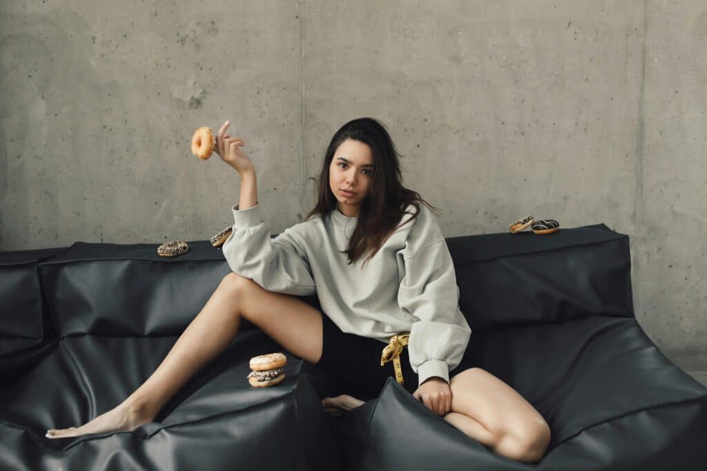 A woman poses with donuts on a comfortable black sofa in a minimalist indoor setting.