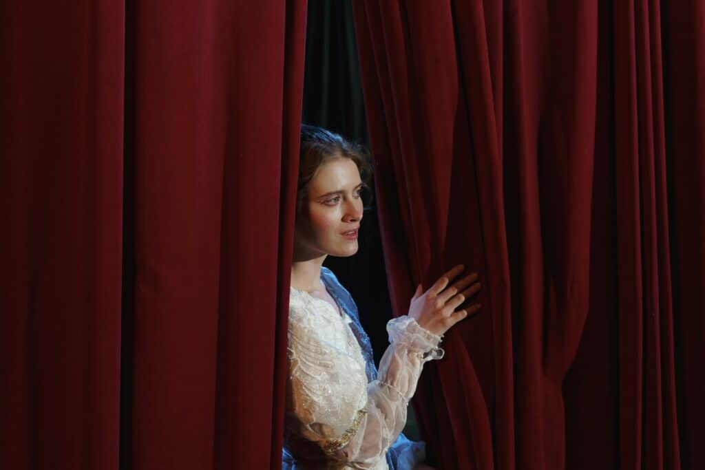 A woman in costume peeks through red theater curtains in a dramatic scene.