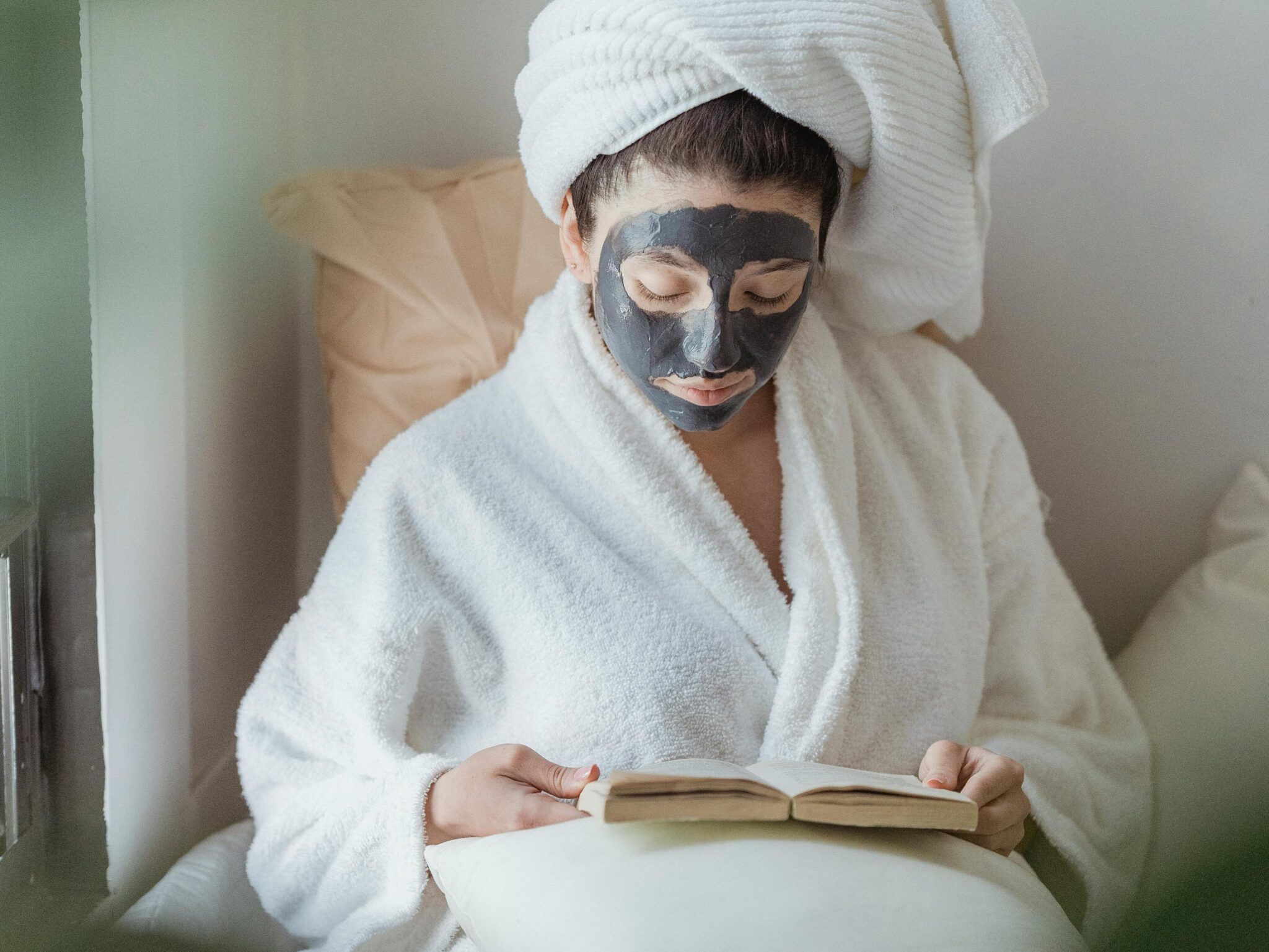 Relaxing self-care moment with a woman in a robe, clay mask, and book, offering tranquility and pampering.