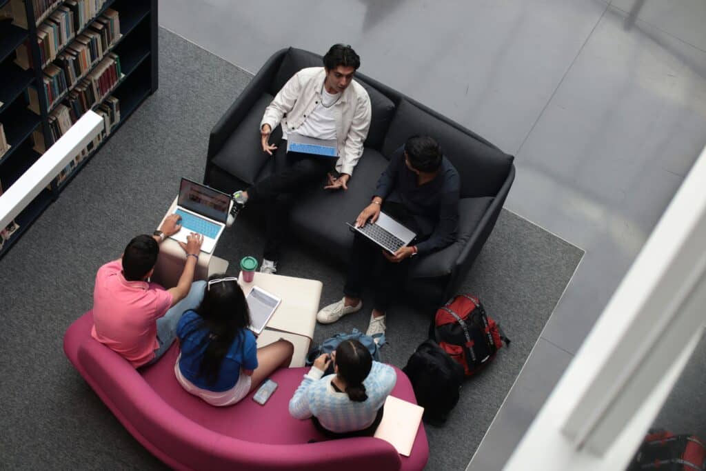 A group of diverse students using laptops and books in a library setting, studying together.